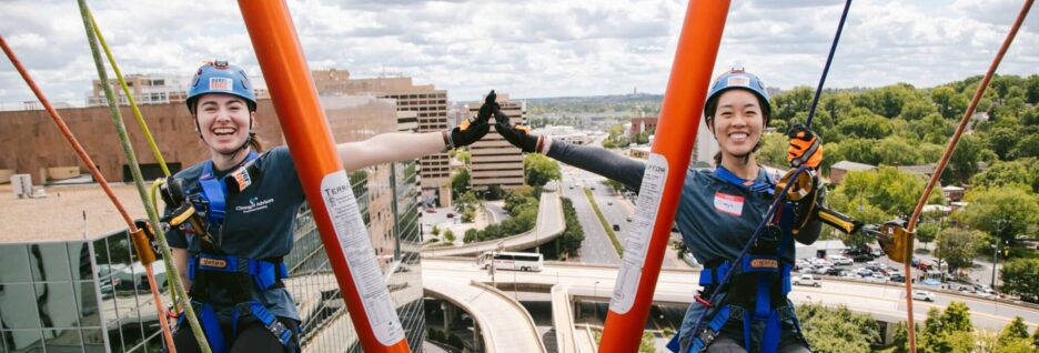 Two women smiling and high-fiving as they rappel down the side of a building.